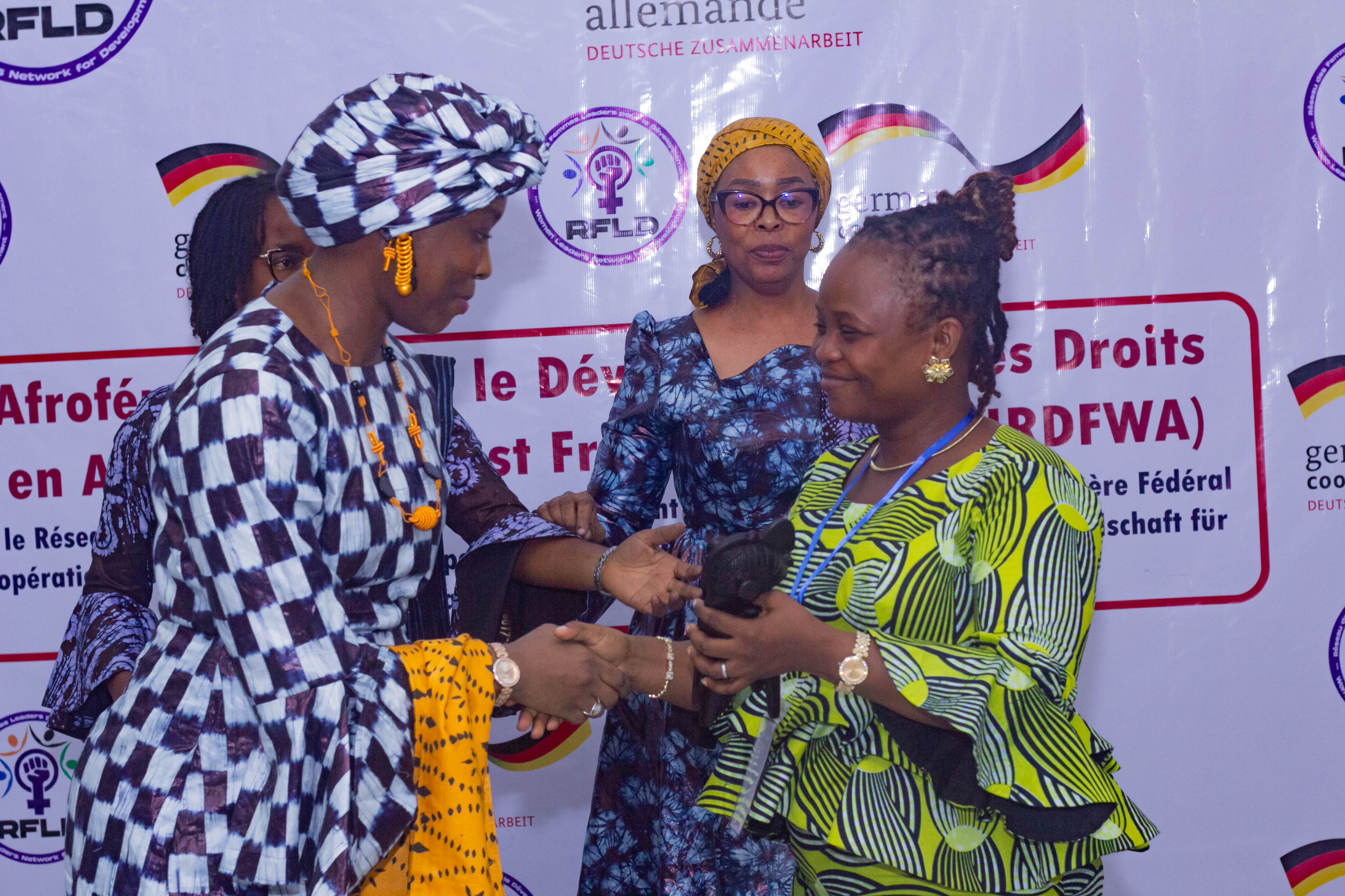 A group of women participating in a formal event where one woman is shaking hands with another, while a third woman observes, all dressed in colorful traditional attire with a background featuring banners related to women's rights and empowerment.