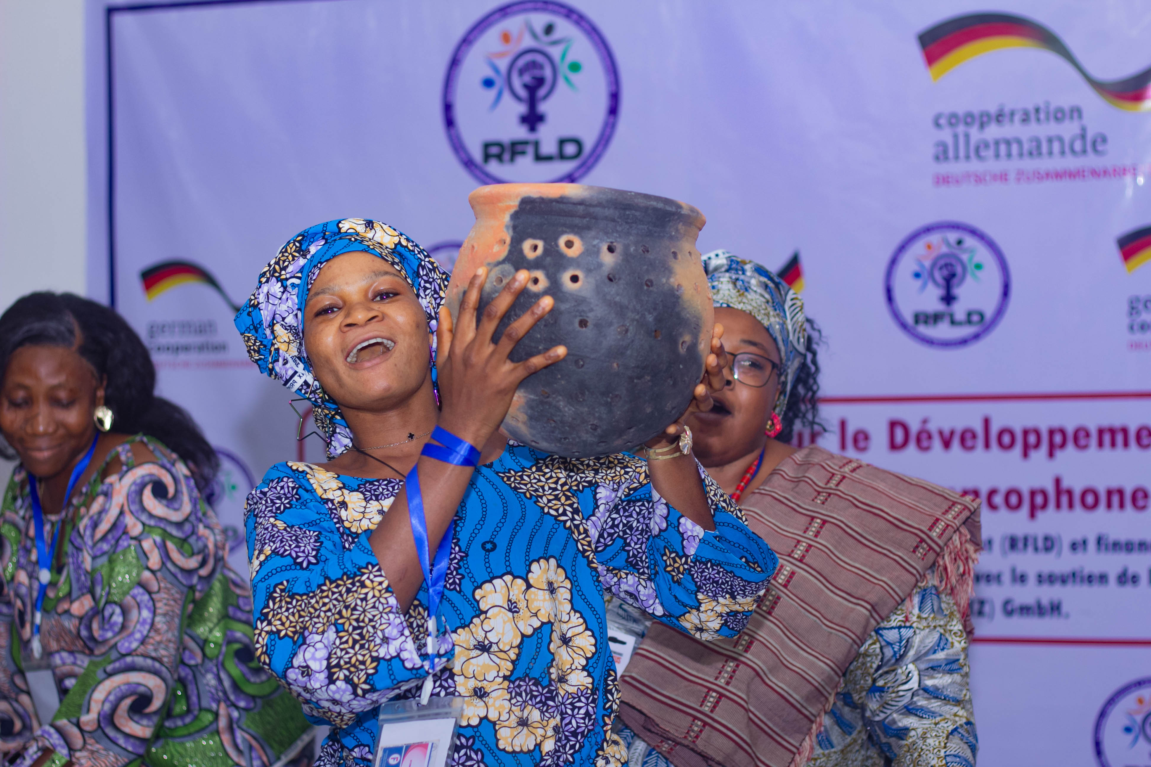 A woman wearing a colorful outfit joyfully holds a traditional pottery vessel at an event, with other attendees in the background.