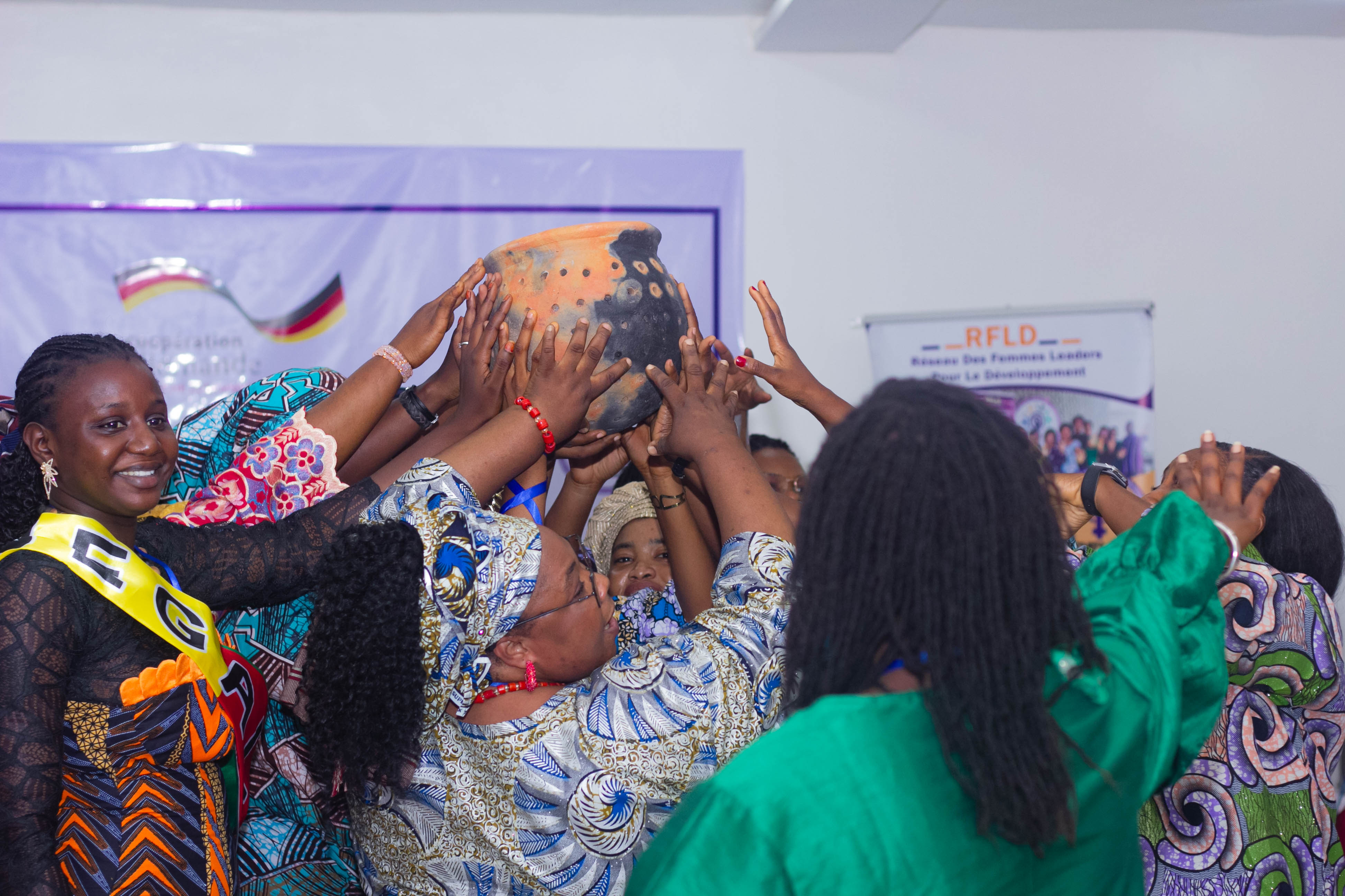 A group of women joyfully participating in a traditional celebration, lifting a decorated pot together, with vibrant clothing and expressions of excitement.