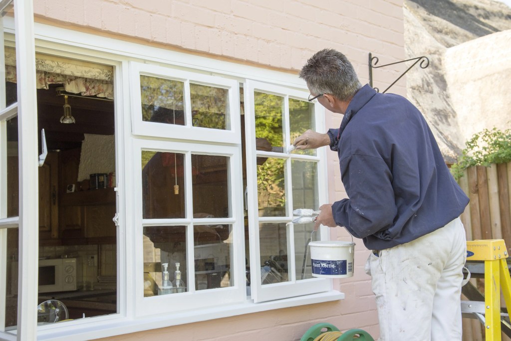 A person painting a window frame with a brush, wearing a blue shirt and white pants, with a paint bucket beside them.