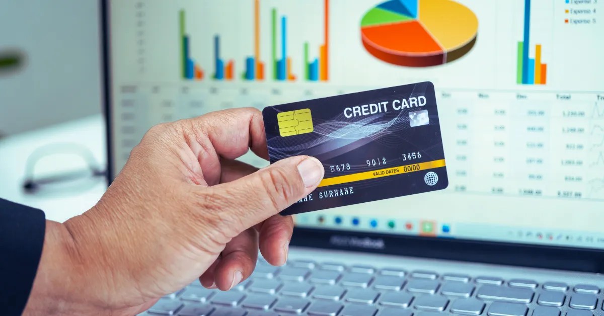 A hand holding a credit card in front of a laptop displaying financial graphs, illustrating strategies to improve credit scores.