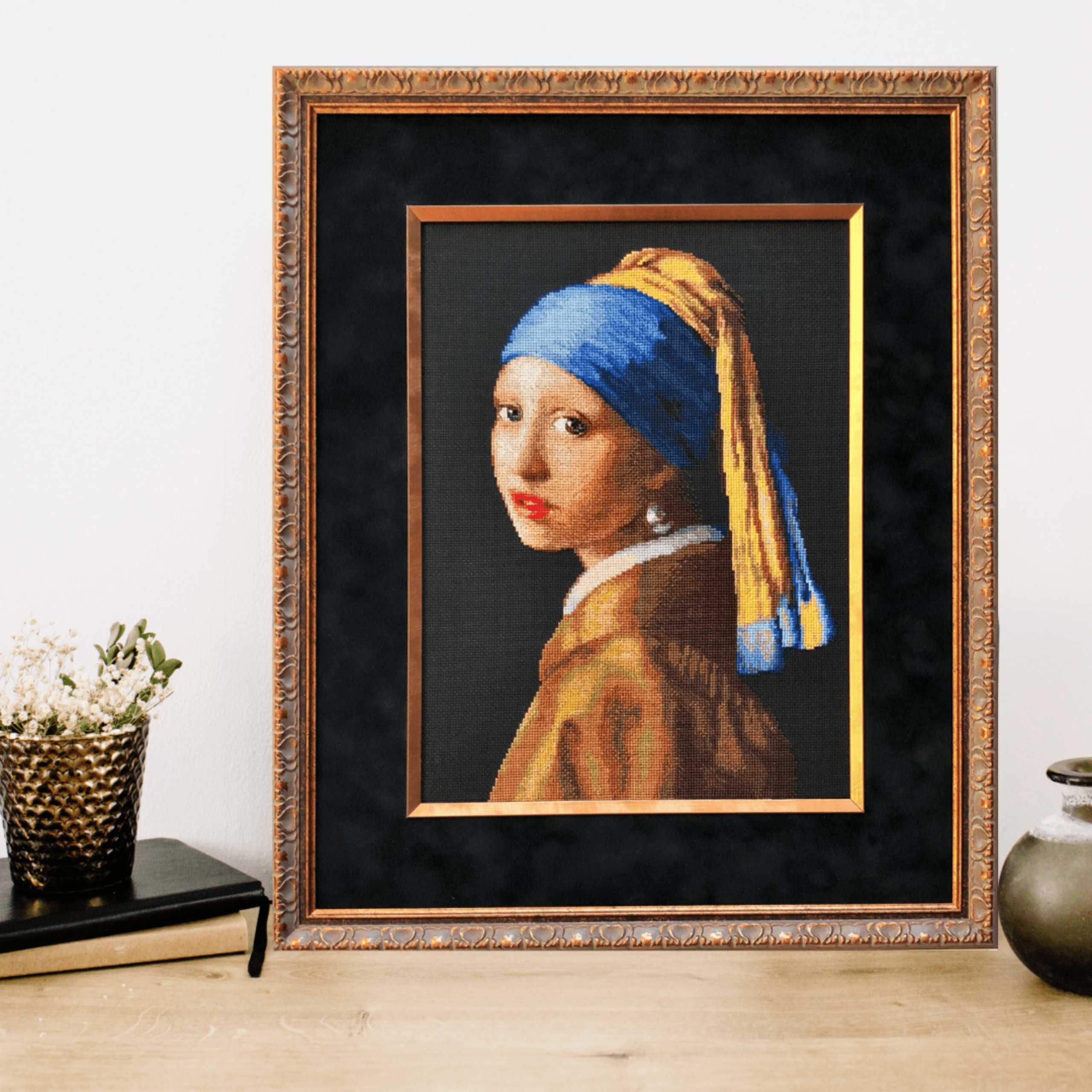 Cross-stitch embroidery of Vermeer’s Girl with a Pearl Earring, featuring a young woman wearing a blue and yellow headscarf and pearl earring. Set against a dark black background in an ornate silver frame. Design from RIOLIS kit 100/063.