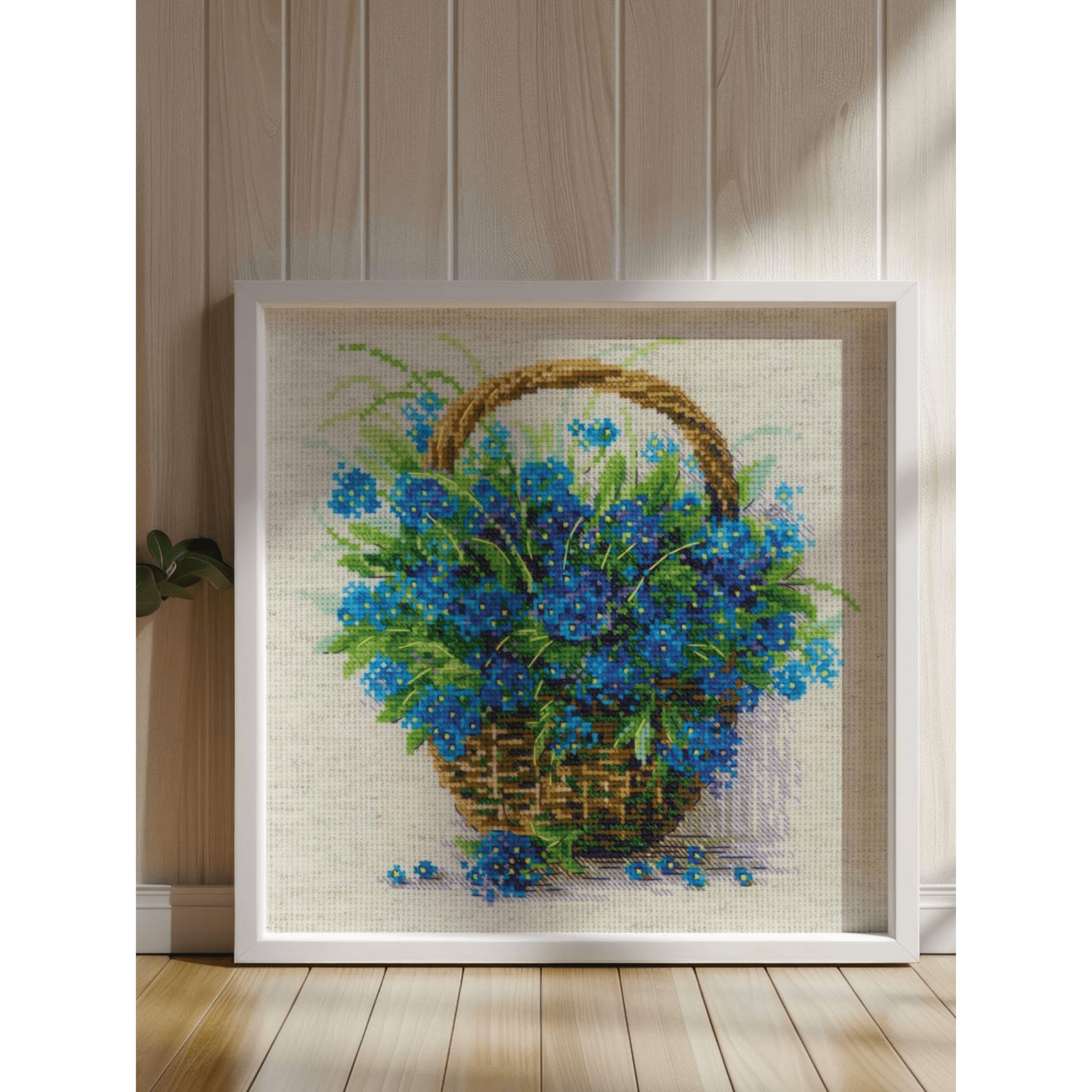 Cross-stitch design of a wicker basket overflowing with vibrant blue forget-me-not flowers with scattered petals around it.