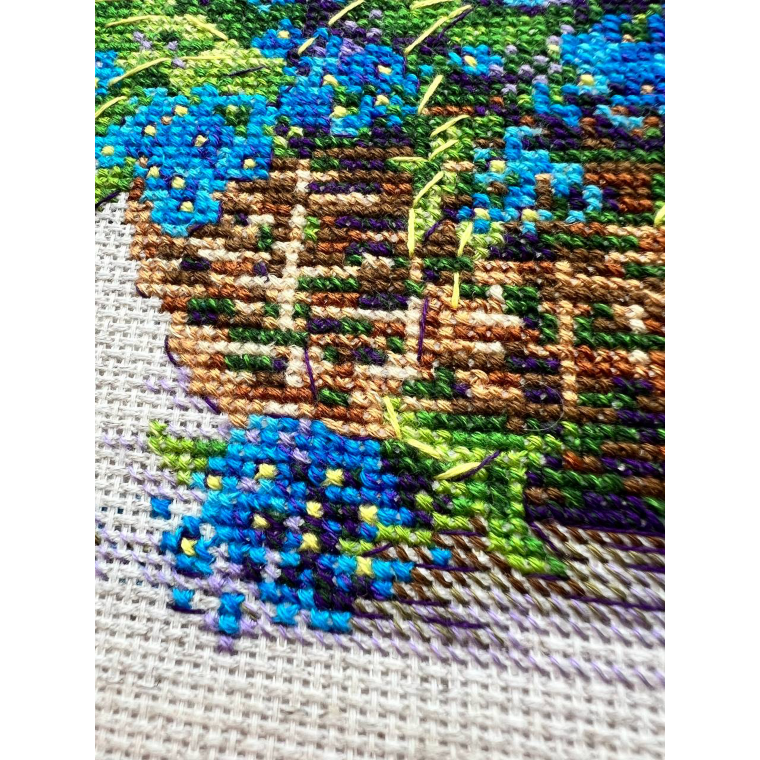 Cross-stitch design of a wicker basket overflowing with vibrant blue forget-me-not flowers with scattered petals around it.
