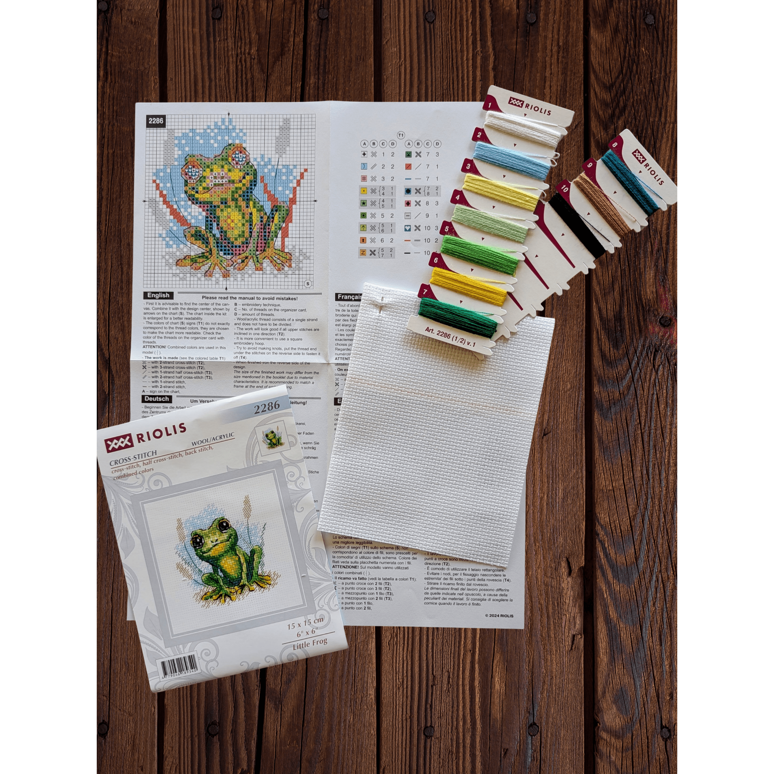 Cross-stitch design of a small green frog with bright yellow accents sitting in grass with a watercolor-style background.