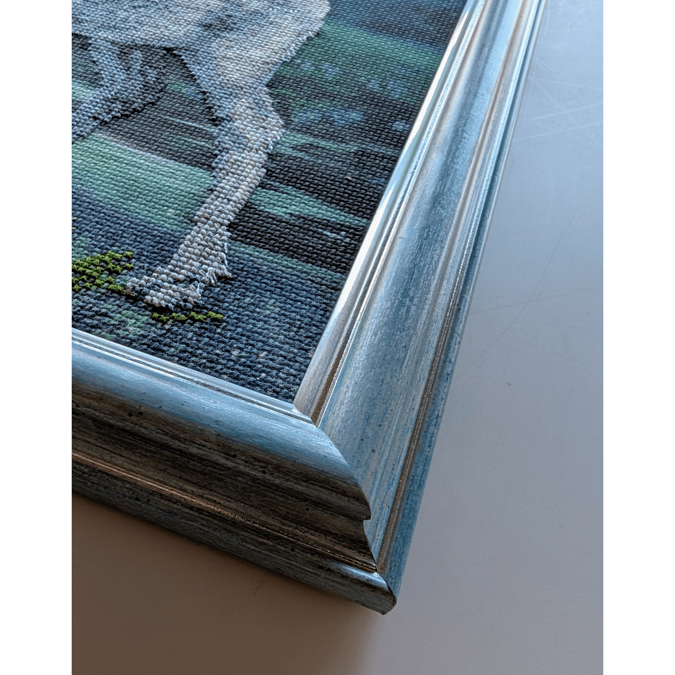 Framed cross stitch artwork of a white stag with leafy antlers standing by a lake under a glowing northern lights sky at night.