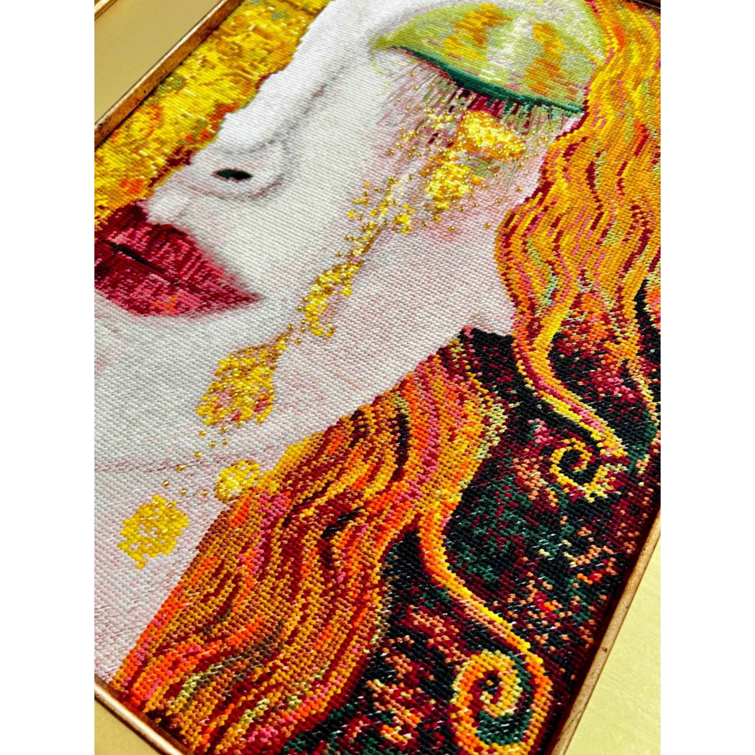 Cross-stitched portrait of a woman with flowing auburn hair, green eyeshadow, and golden tears against a richly textured gold background.