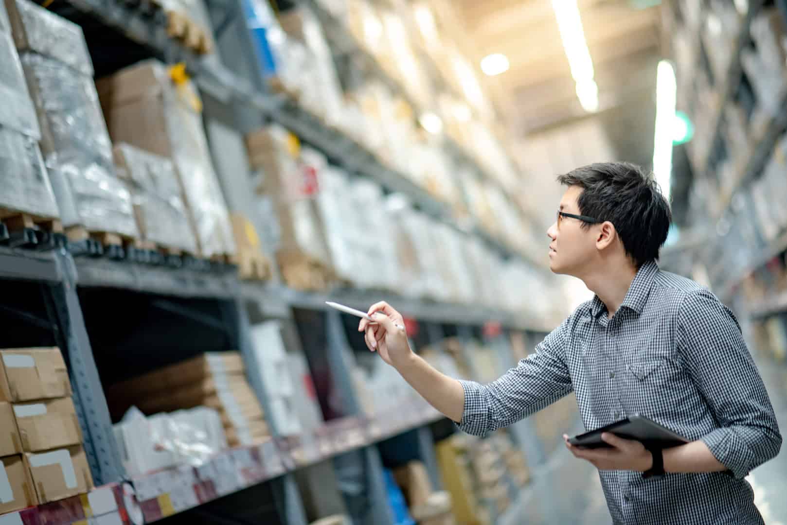 A warehouse worker conducting a physical inventory count, holding a tablet and pointing at stock items on high shelves in a well-organized warehouse.