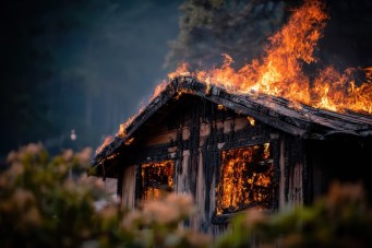A wooden cabin is engulfed in bright flames, with smoke rising into the air, surrounded by blurred foliage and trees in the background