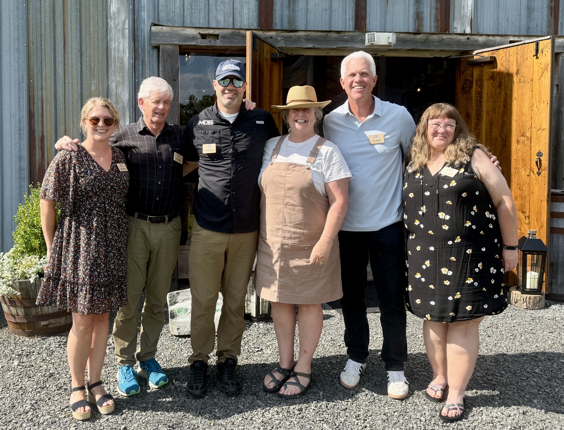 Six adults stand side by side, smiling in front of a rustic wooden building with open doors, on a sunny day—a place that exudes history and evokes the enduring spirit of the Mission.