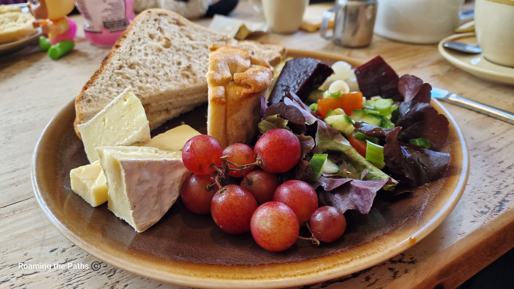 This image shows a traditional Ploughman’s Lunch served on a wooden board. The meal consists of a variety of classic components, including a generous wedge of cheddar cheese, thick slices of crusty bread, a portion of butter, and a selection of pickles and chutneys. Fresh salad items such as lettuce, cucumber, and tomatoes are also arranged on the board, alongside a sliced apple and boiled egg. The presentation is rustic and hearty, evoking the classic British countryside meal, perfect for a simple yet satisfying lunch.