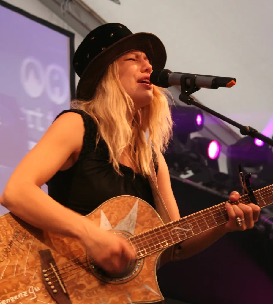 Blonde Sängerin mit Hut singt auf Bühne und spielt Gitarre