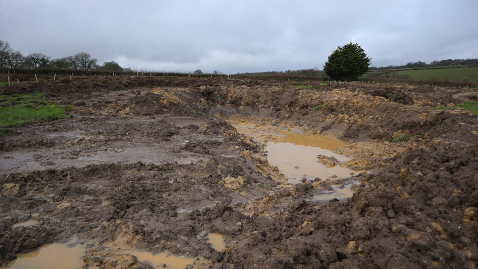 a very recently dug pond, with clay in the base and some water. It is surrounded by mud and very little grass.