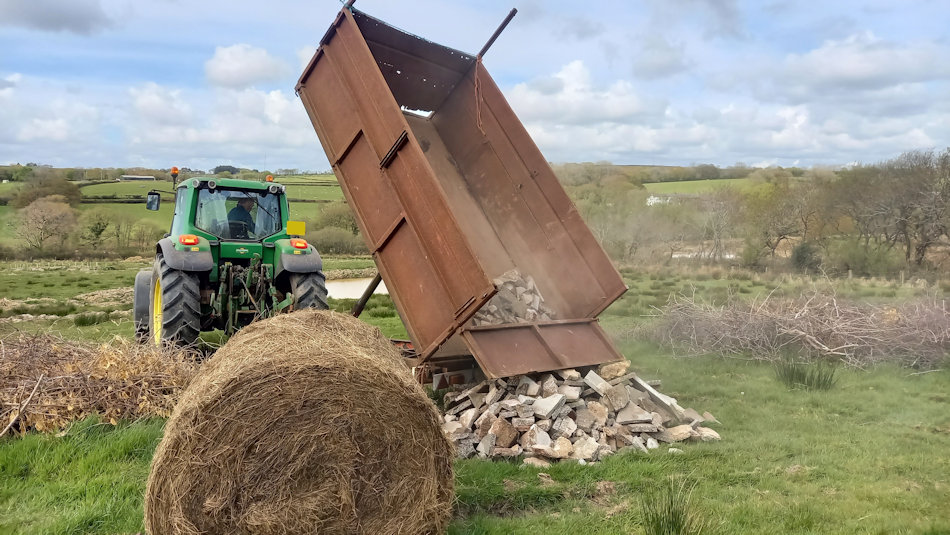 rubble being emptied out of a trailer, behind an old bale and between piles of brash