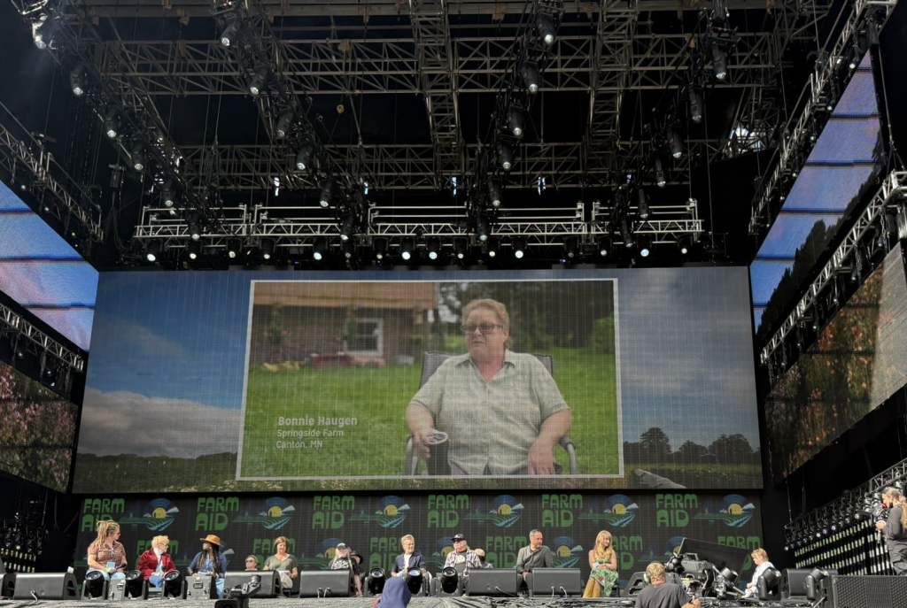 PHOTO 1 092025 Farm Aid Press Conference Bonnie Haugen big screen A large screen behind a stage lined with people in chairs shows a womean sitting in a folding chair in her grassy yard.