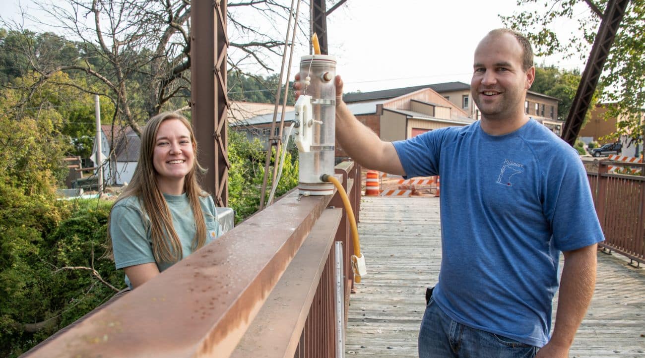 A young woman (left) and a young man (right) stand on either side of a wide, rusted bridge railing and hold a container of water with a rubber tube coming out of the bottom of the container.