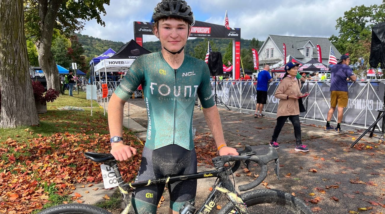 A young man wearing a bike helmet and a green and black one-piece biking uniform splattered in mud stands behind his bike with his hand on the seat and handlebars.