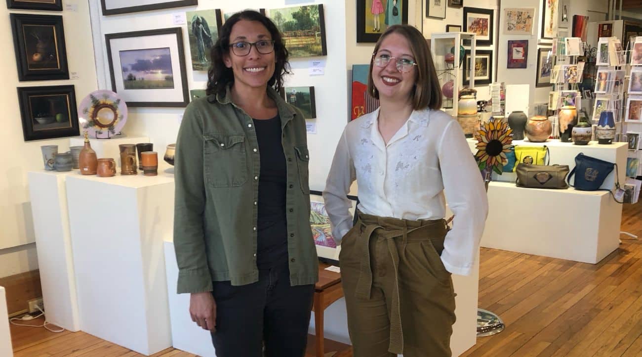 Two women stand in an art studio with pottery and paintings in the background.