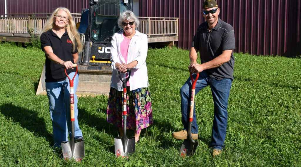 PHOTO 1 Spring Valley Ground Breaking From left to right: two women and a man hold shovels in a grassy area in front of a wooden deck attached to a metal building wall.