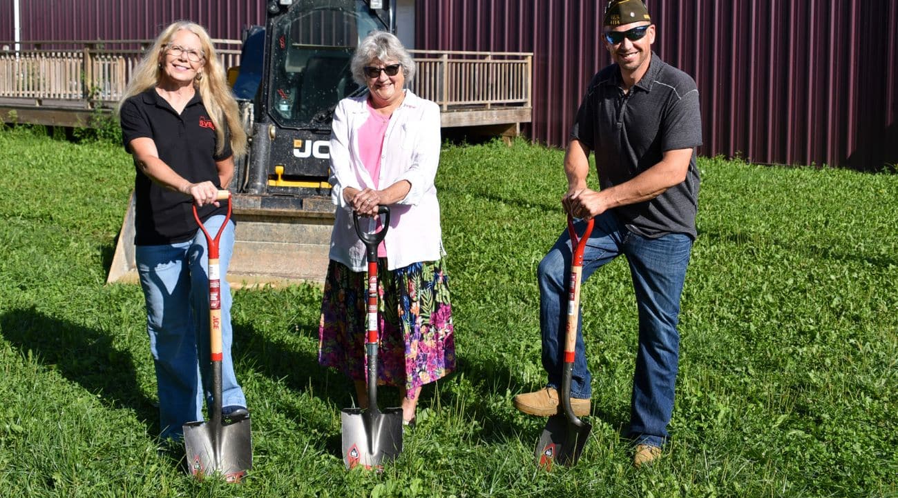 From left to right: two women and a man hold shovels in a grassy area in front of a wooden deck attached to a metal building wall.