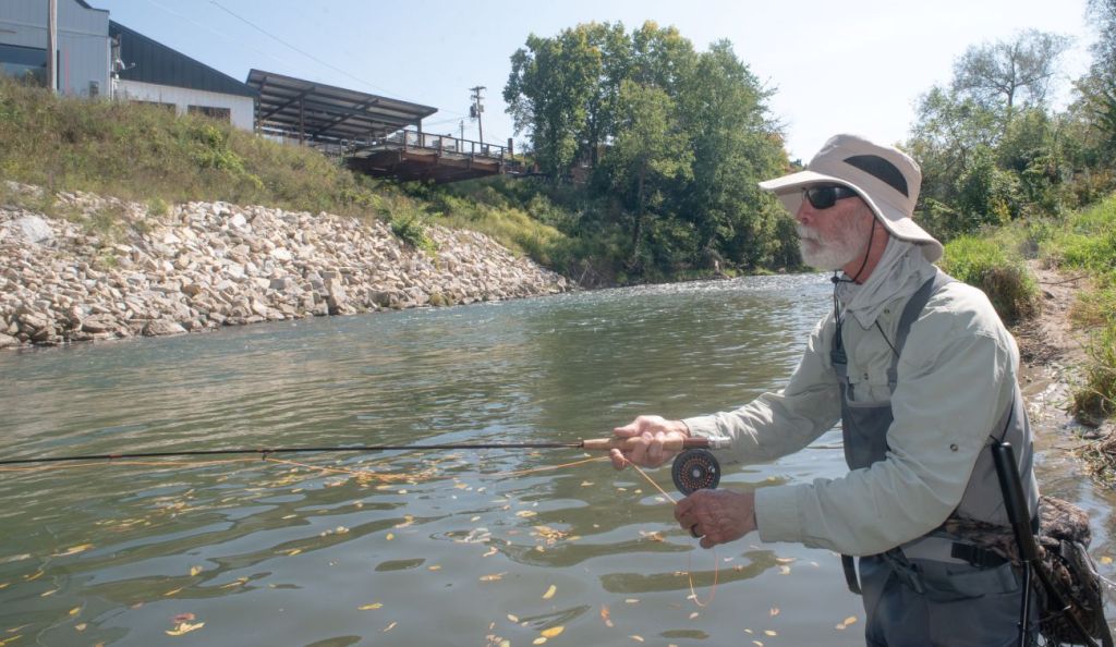 And older man with a long sleeve shirt, wide brim tan hat and sunglasses holds a fishing pole while standing in the river. Behind him is a large rock wall of riprap.