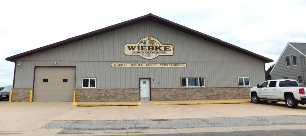 A large pole shed type building with taupe siding and a cement parking lot in front. The sign on the building reads Weibke.