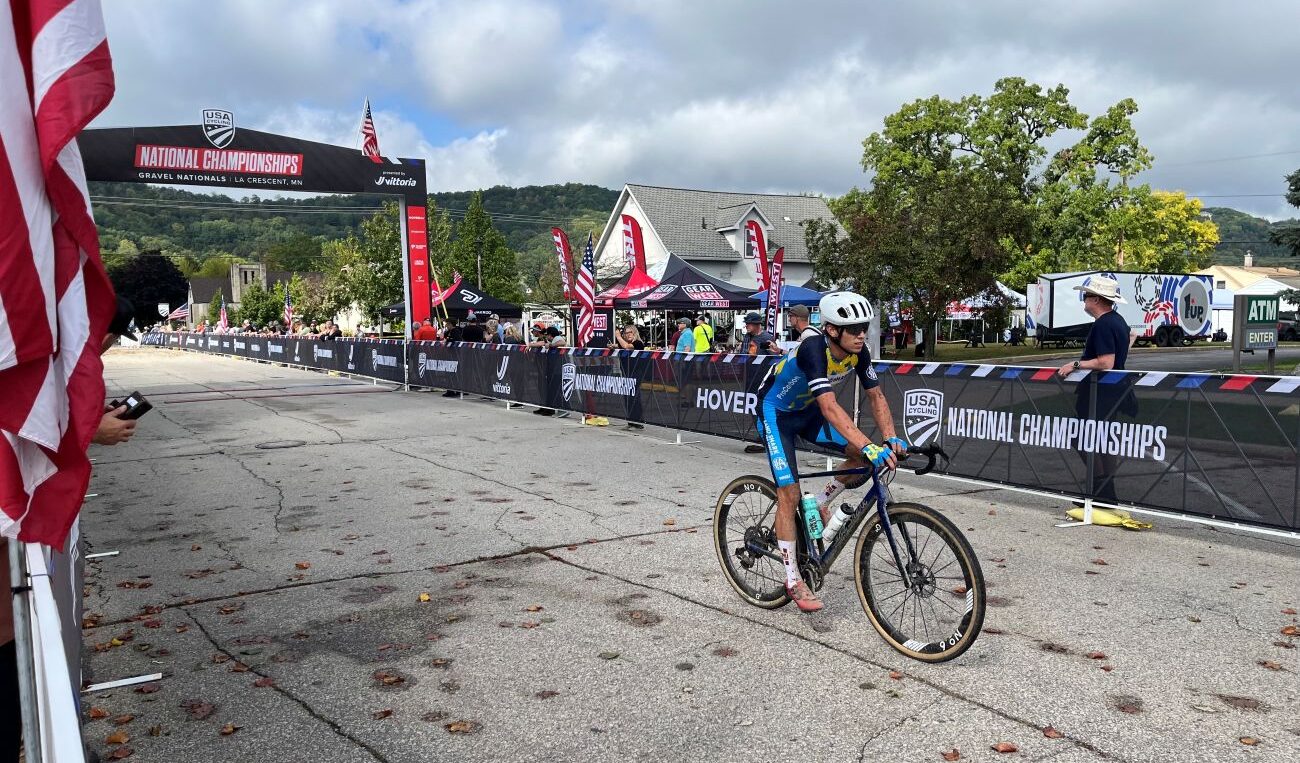 A biker cycles through a small crowd behind fences lining the finish area of a bike race. On the upper left corner is an archway marking the finish line.