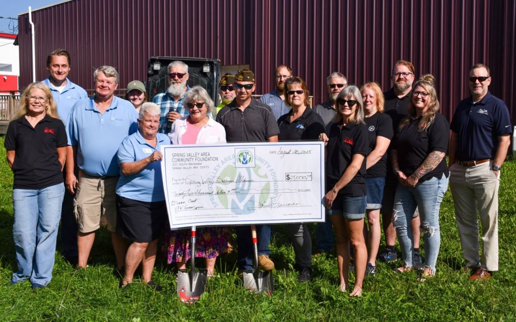 A large group of a field holding an oversized check stand in a grassy lawn in front of a large shed with dark burgundy metal siding.