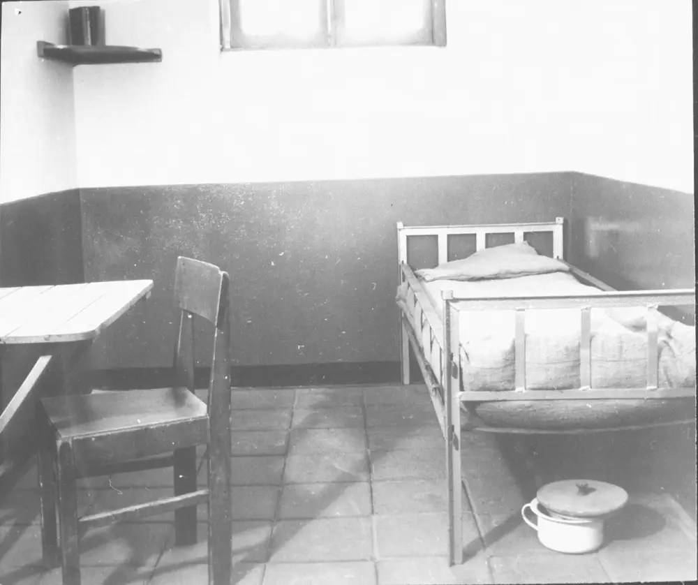 Black-and-white photograph of a prison cell in Kamp Vught, showing a sparse wooden bed with a thin blanket, a chair beside a small table, and a chamber pot beneath the bed. Light enters from a high window.