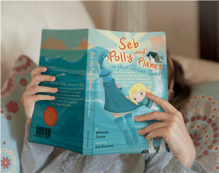 Child holding open "Seb Polly and the Fair Ocean Quest" children's book with blue cover illustration