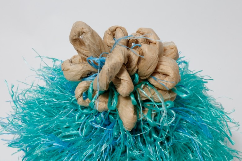 lose-up of knotted tan paper printed with sewing marks, tied with blue strands atop dense turquoise fringe.