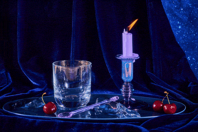 A hand places a chill cube in glass before filling the glass with water. The glass rests on a shining metal plate along with a candle and fresh cherries.