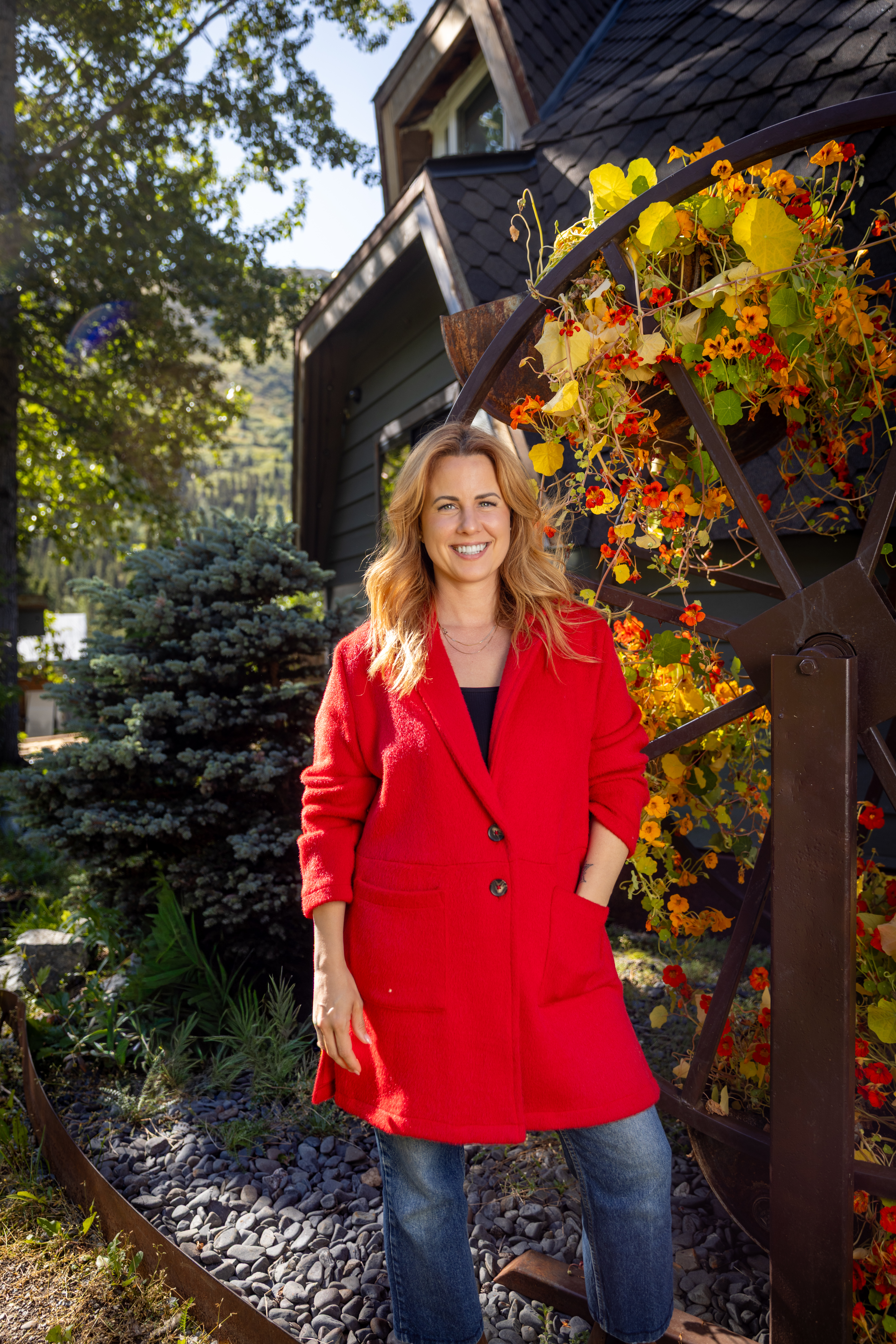 Ruby Jones in a red coat smiling outside a home surrounded by flowering vines