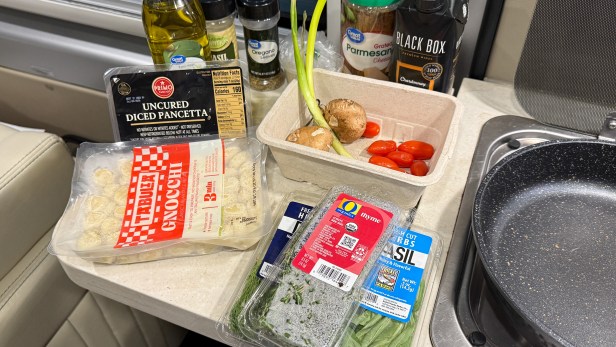 A countertop with various cooking ingredients including uncured diced pancetta, gnocchi, herbs, cherry tomatoes, mushrooms, and cooking oils, arranged next to a frying pan.