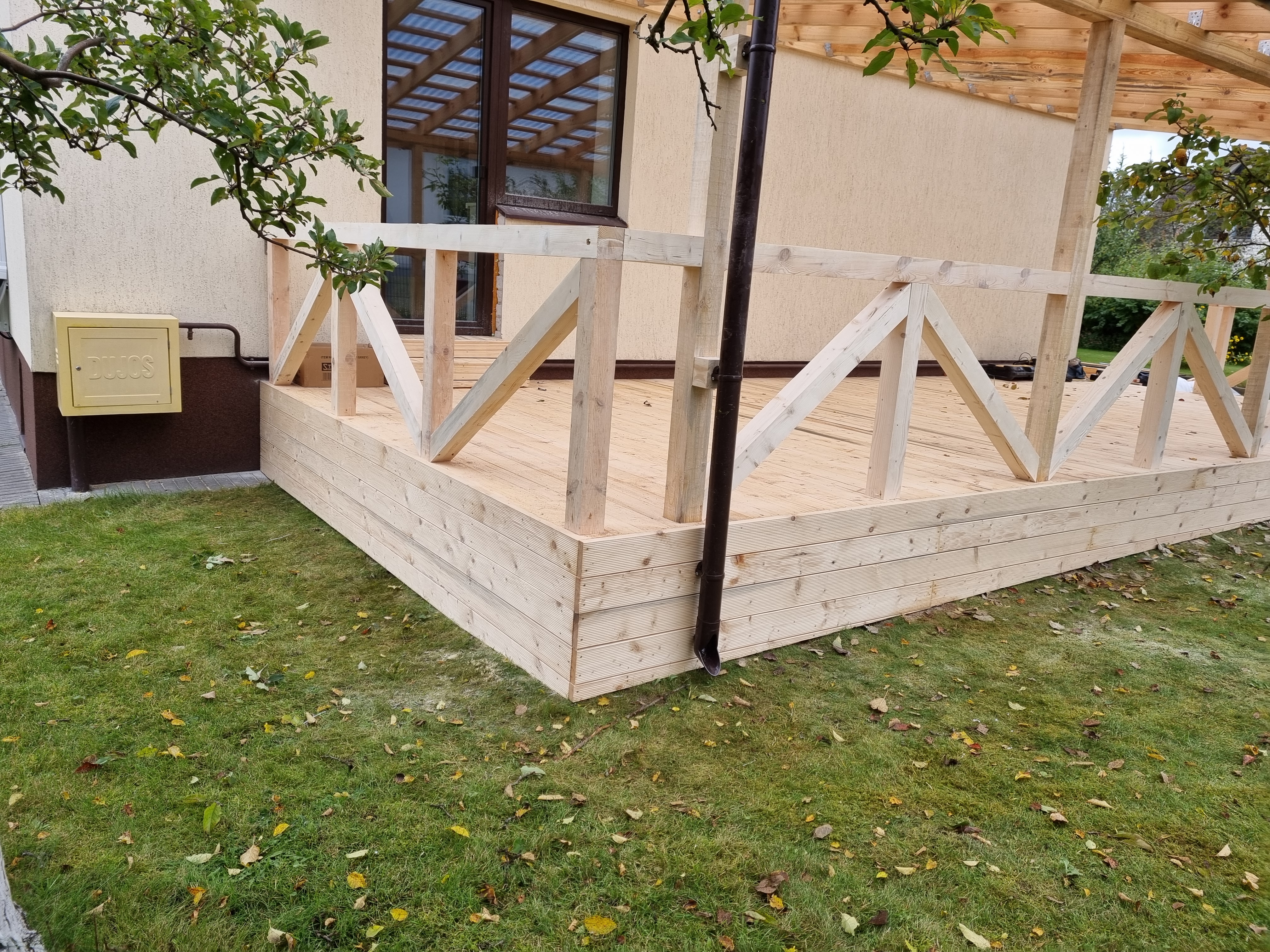 A wooden deck is being built next to an exterior wall of a cream-colored house with a floor-length, tinted glass window. I...