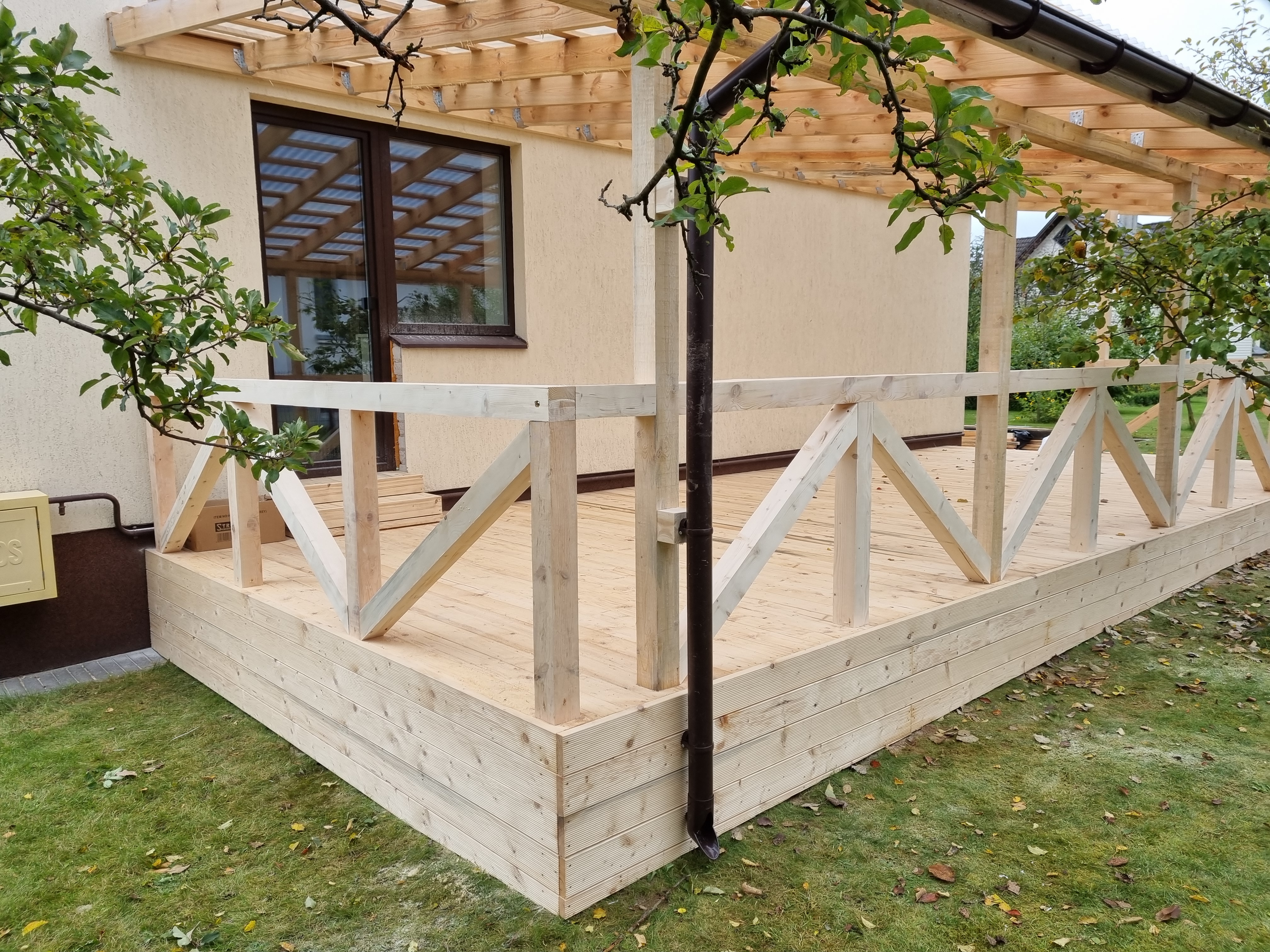 An outdoor wood deck with a low protective railing and gutter under a wooden roof is being built on a grass lawn in front ...