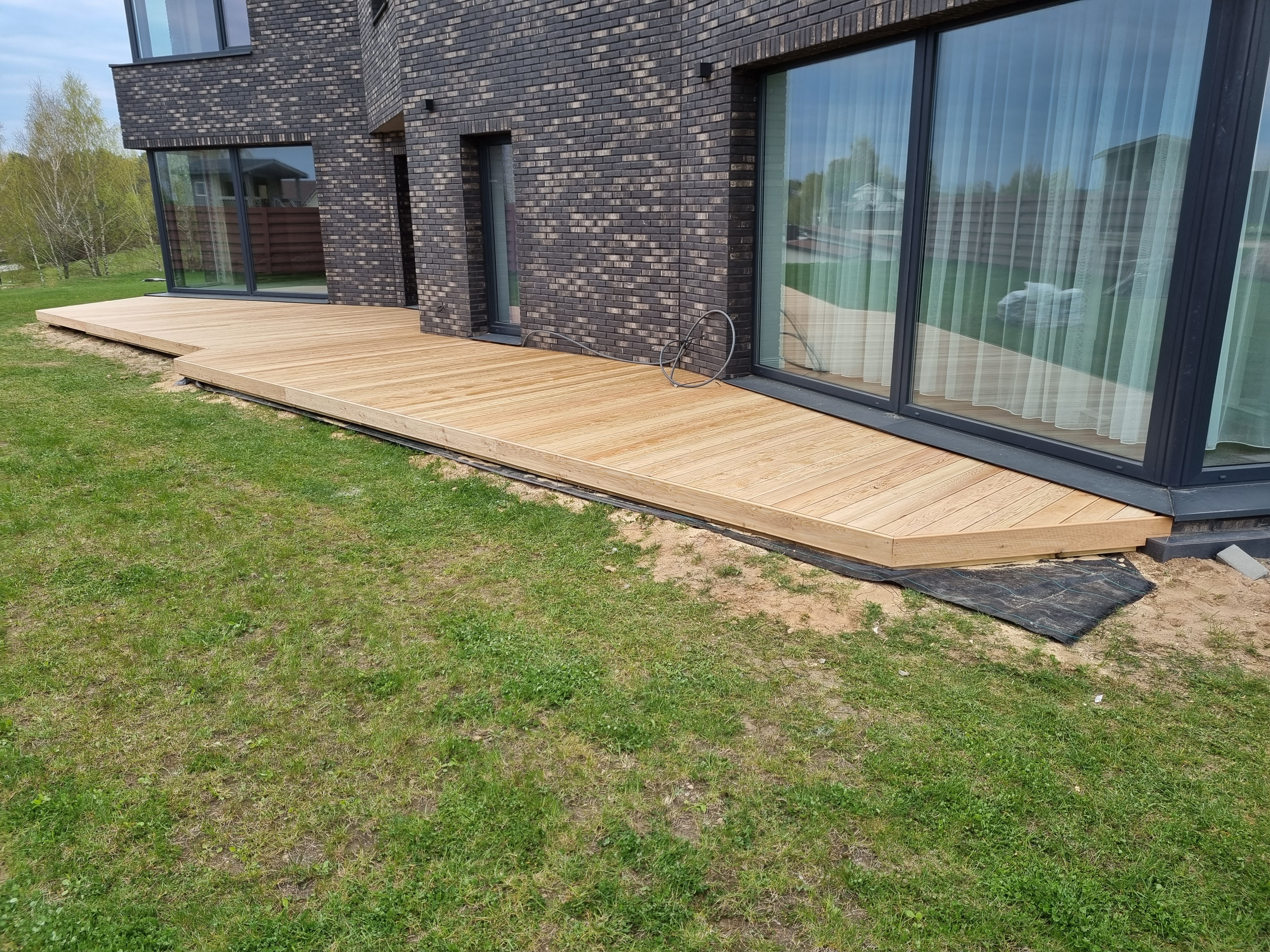 A modern, dark brick house with a flat deck at the patio covered in sunshine, which makes the grass underneath appear slig...