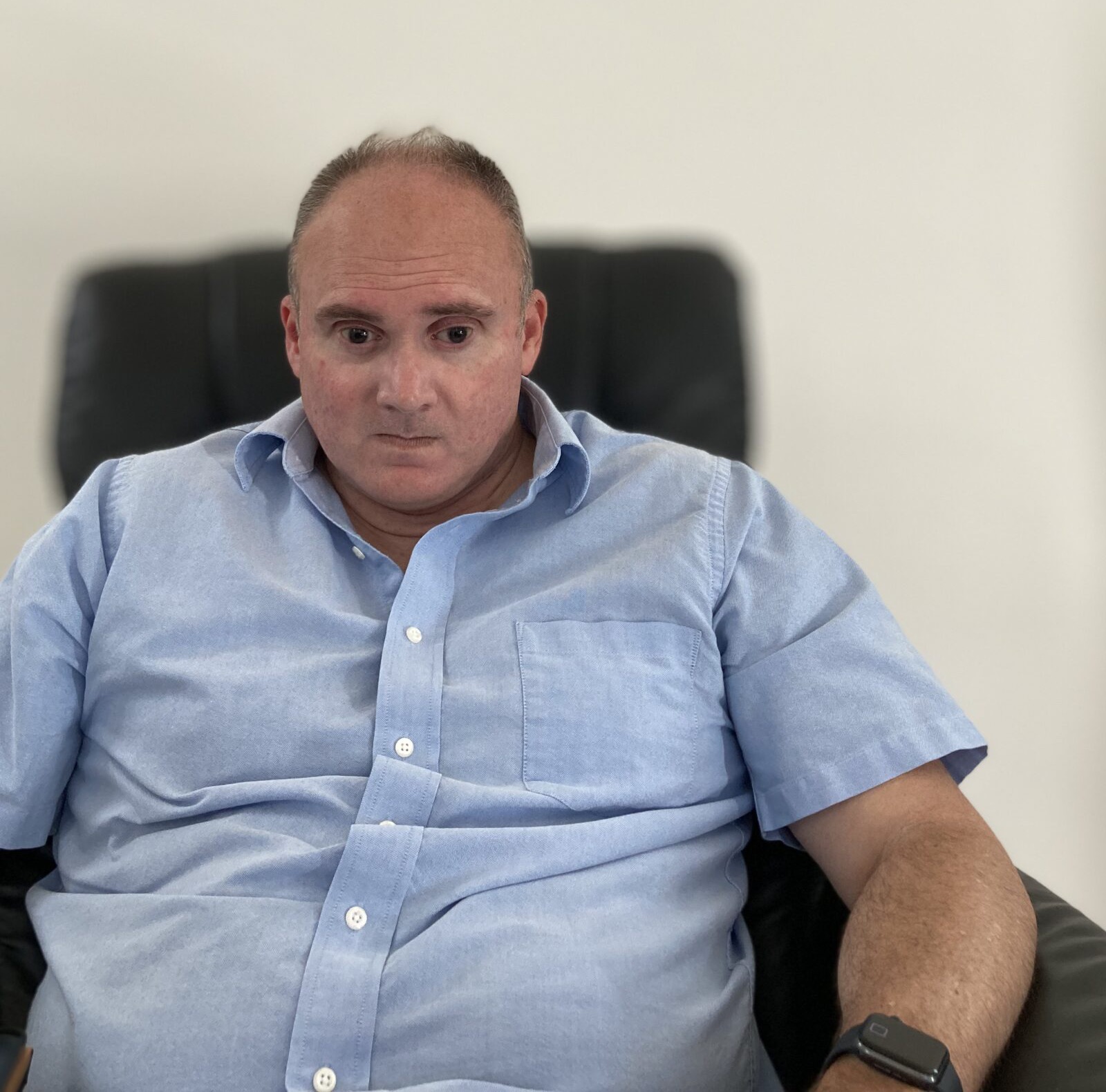 Ryan Hearn seated in a black leather office chair against a plain wall, wearing a light blue button-down shirt, looking slightly away from the camera with a neutral expression.