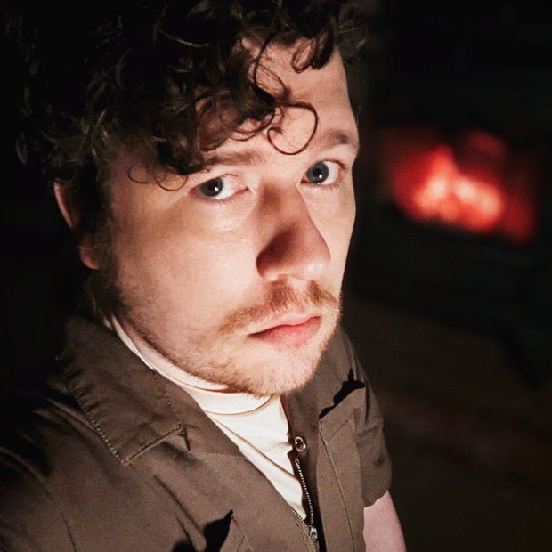 I'm wearing a brown jumpsuit, looking at the camera, with a wood stove burning in the background.