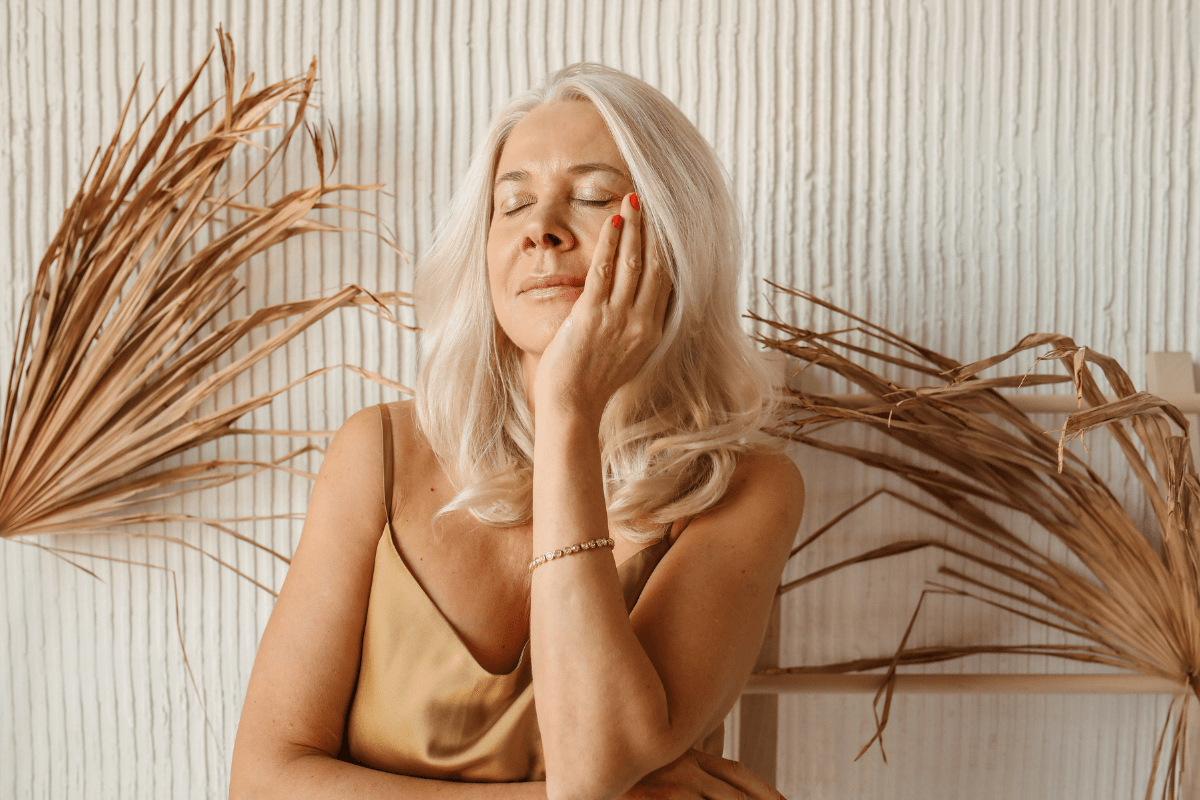 A mature woman with blonde hair resting her hand on her cheek with eyes closed, illustrating a quick self care skincare routine for healthy aging.