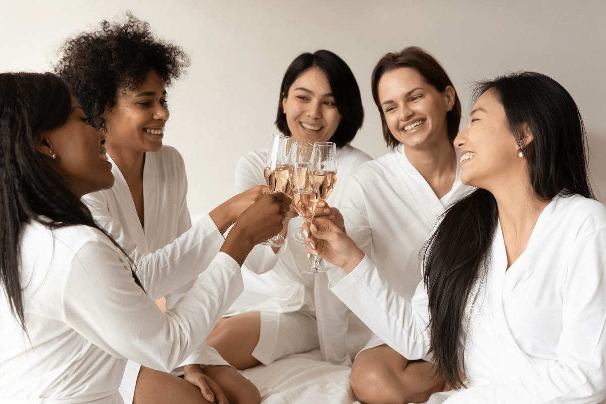 A close-up shot of five diverse women in white spa robes, smiling and clinking champagne glasses together in a toast