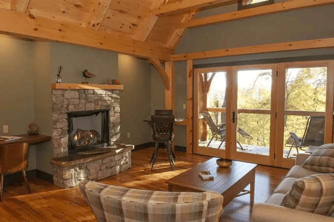 Cozy living room featuring a stone fireplace with a wooden mantle, plaid chairs, and a coffee table beside large glass doors to a deck.