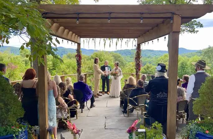 Outdoor wedding ceremony on a wooden pergola overlooking a green valley, couple exchanging vows as guests sit and watch.