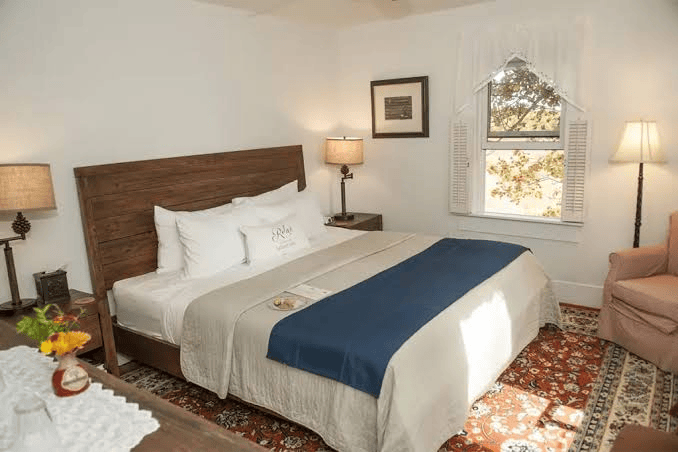 Cozy hotel bedroom with a large wooden bed, white linens, blue bed runner, and two nightstands with lamps on either side of the windowed wall.