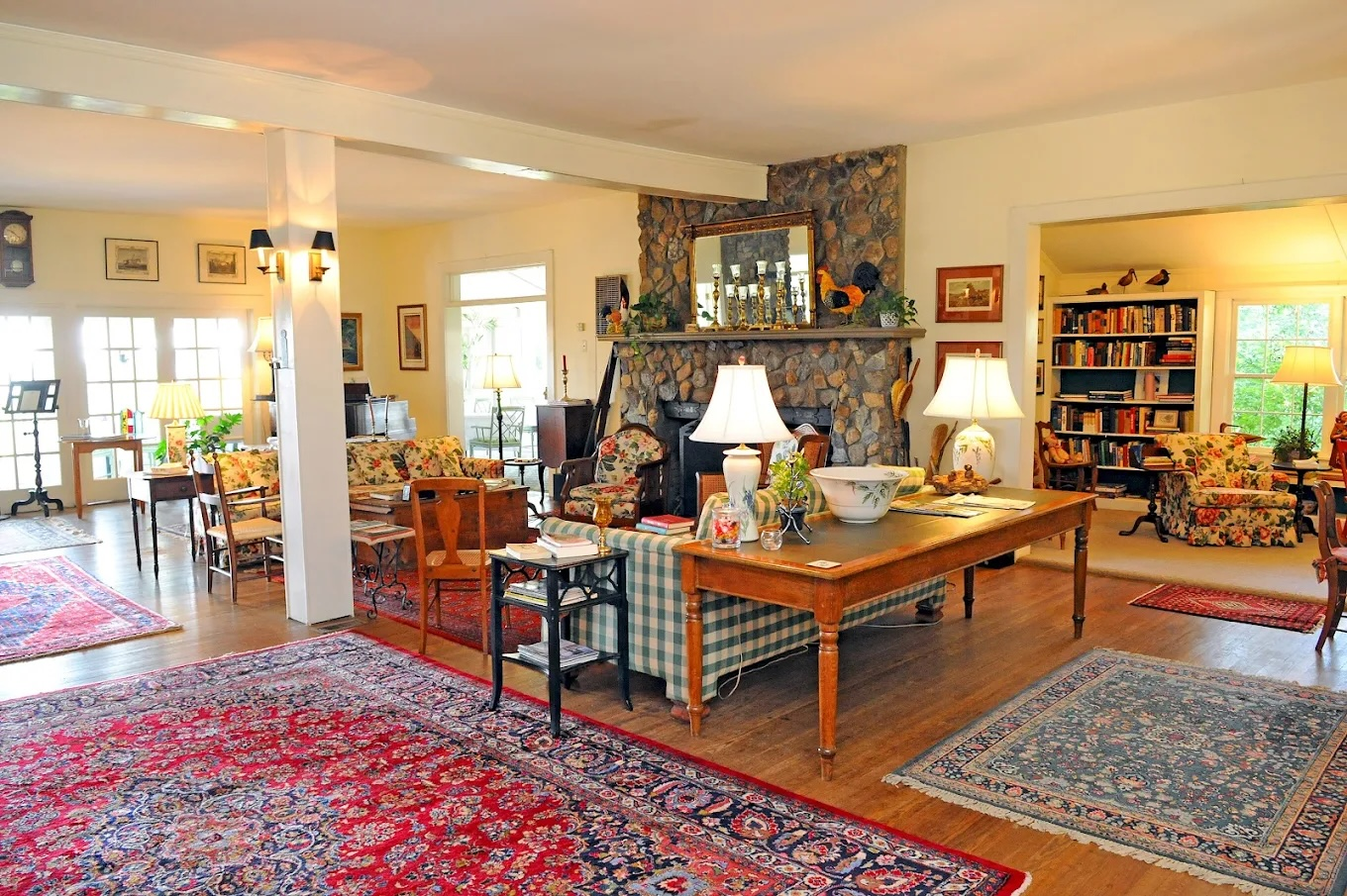 Cozy living room with a stone fireplace, multiple sofas, lamps, and a large bookcase in the background.
