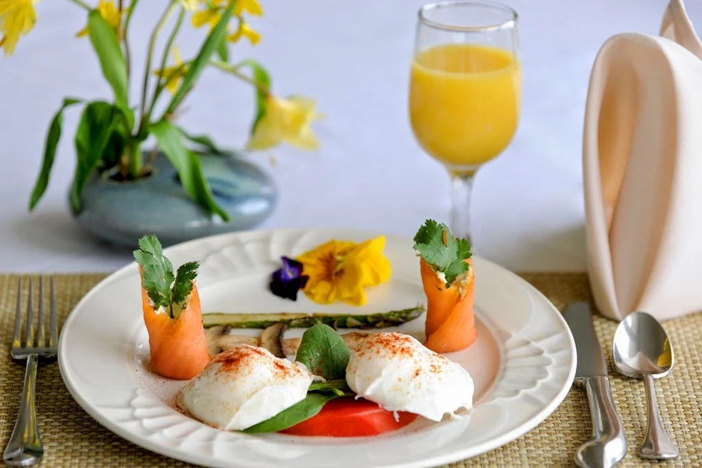 Elegant breakfast plate with poached eggs, asparagus, and carrot roulades, garnished with greens and a tomato slice; glass of orange juice in the background.