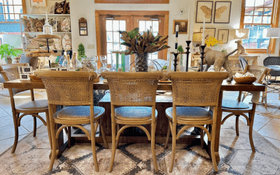 Long wooden dining table with wicker chairs in a bright showroom, topped with plants and decorative objects.