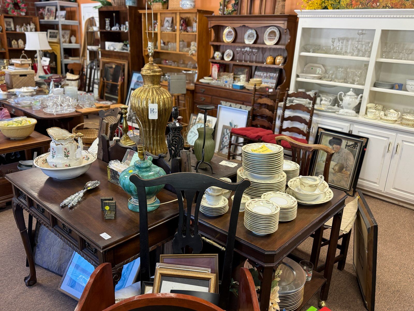 A cluttered antique shop with a dark wood table piled high with stacked china, a floral pitcher, glassware, and decorative metal lamps, shelves of porcelain and frames in the background.