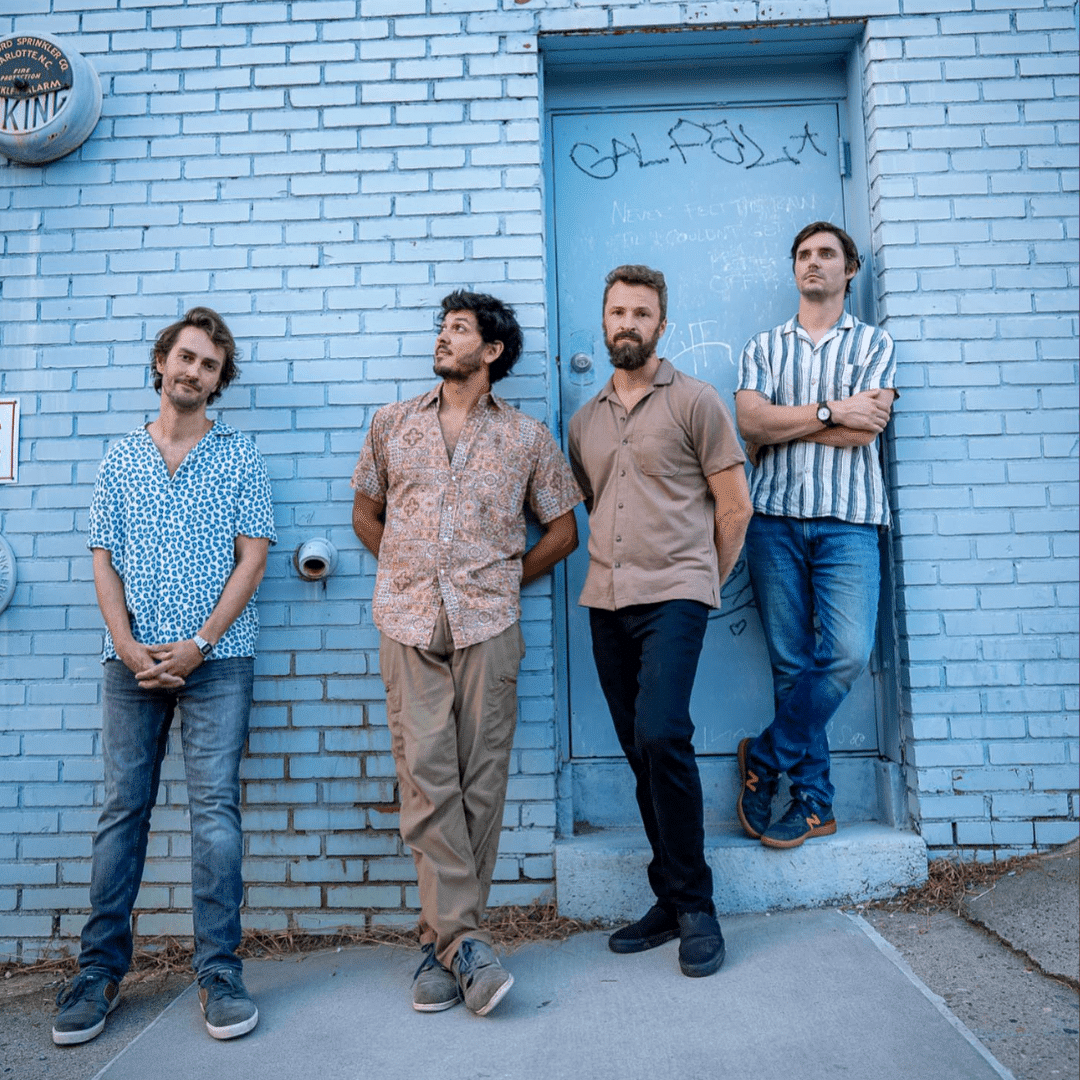 Four casually dressed men leaning against a blue brick wall beside a graffiti-covered door.