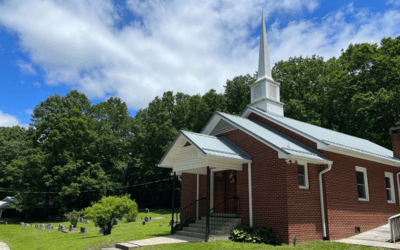 Brick church with white trim and a tall steeple on a grassy lawn, with trees and a small cemetery visible to the left.